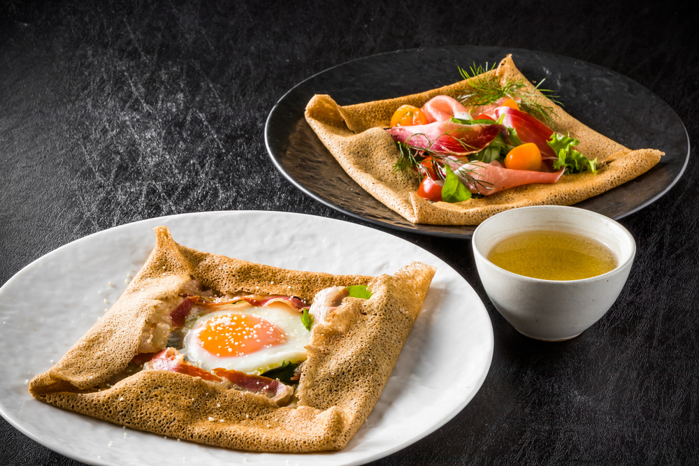 Two buckwheat crepes, one with an egg, and one with smoked meat and greens, sit on separate plates, like those made at Breizh Pan Crepes in Charleston.