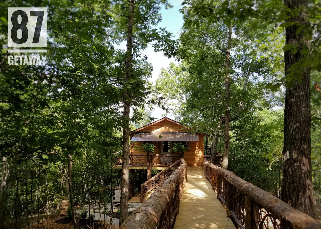 A wooden treehouse cabin set back in the forest, with a wooden bridge leading up to it. 