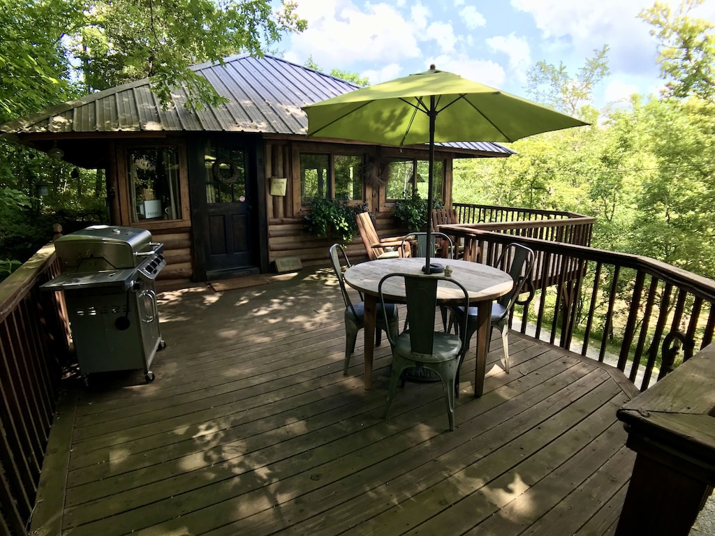 A wooden deck with a wooden cabin, outdoor dining setting and barbecue, with a balustrade overlooking a river and forest. 