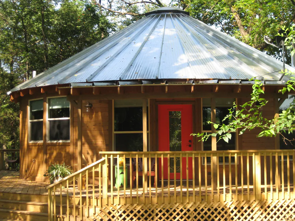 A yurt (round wooden cabin) with a silver pointed roof and red door. An example of glamping in Arkansas. 
