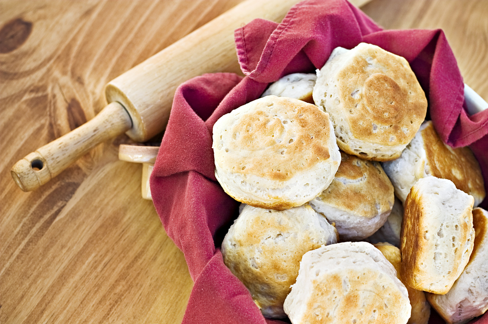 A fresh warm basket of southern buttermilk biscuits. A red cloth and rolling pin accompany the biscuits. 