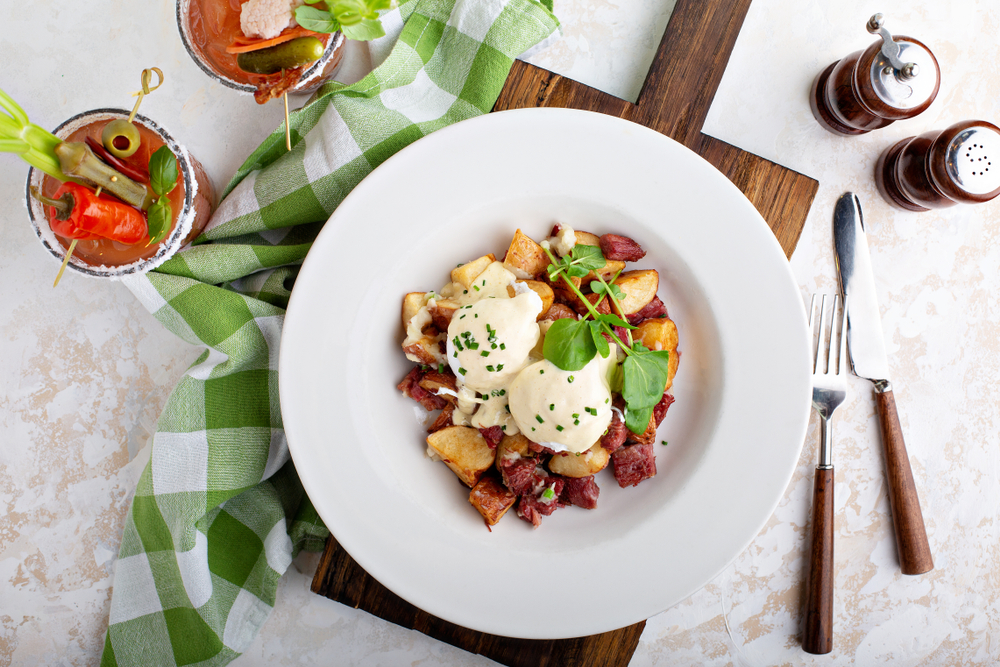 A plate of corned beef hash topped with poached eggs and hollandaise on a table with a green and white checked tablecloth. 