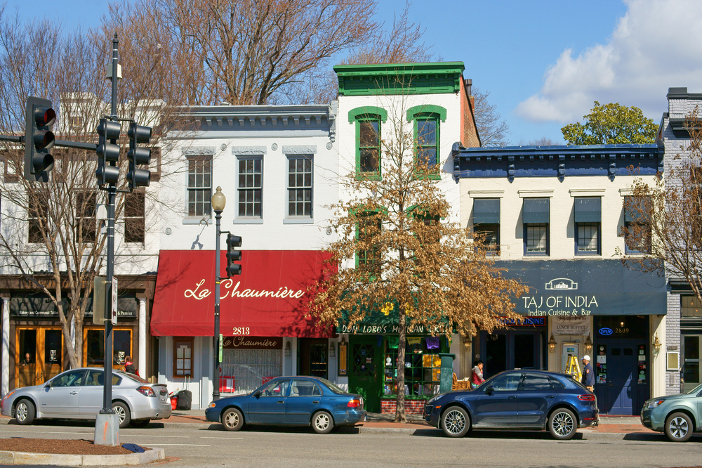 A streetscape of Georgetown DC with cars parked in front of a row of restaurants and shops. 