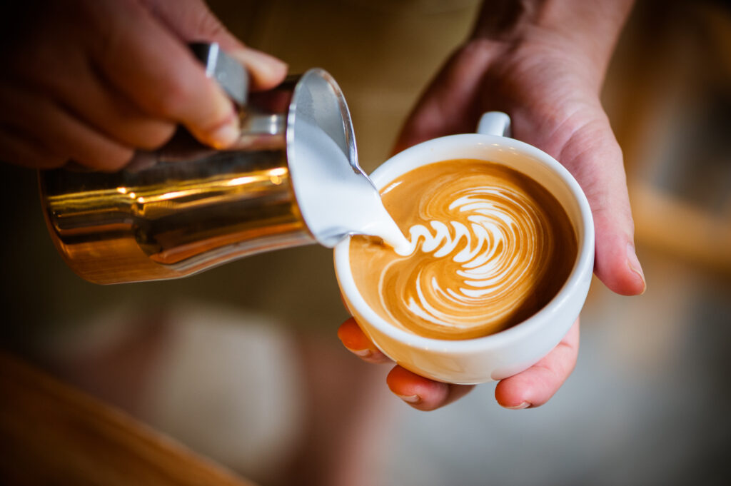 A barista's hands hold a small white cup and a cup of milk. The milk is being poured into the coffee to make latte art.