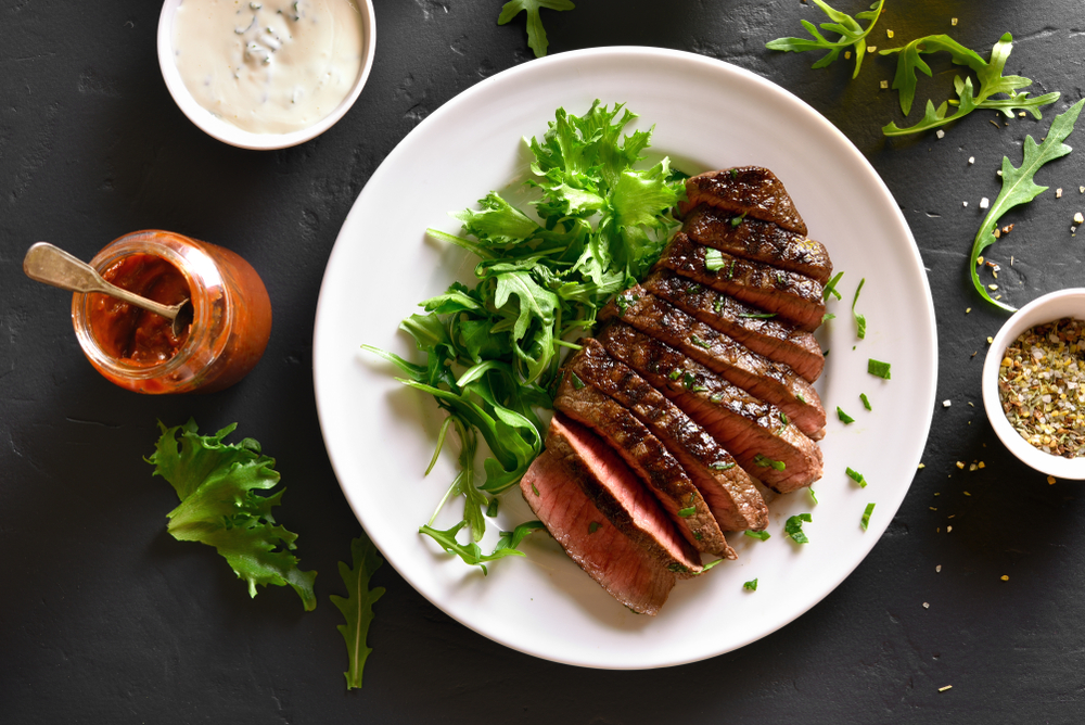 A medium-rare steak sits on a white plate next to a green salad on a black stone table.