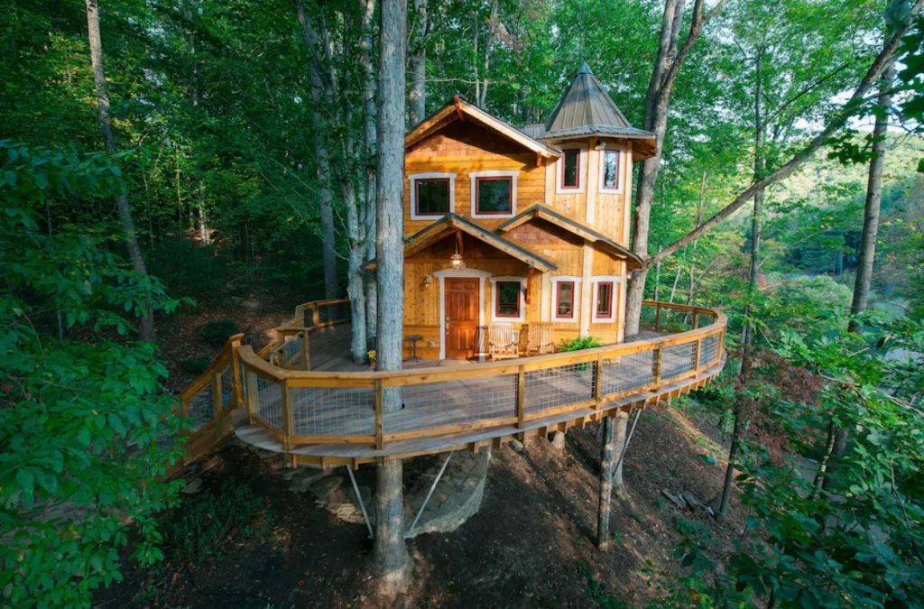 The exterior of a multi-level treehouse with a deck wrapping around it. It is surrounded by trees. A cool spot for glamping in North Carolina.