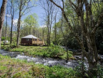 A yurt on the edge of a river in the middle of the woods on a sunny day. It's a great spot for glamping in North Carolina.