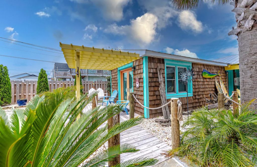 The exterior of a small beach bungalow. It has cedar siding, blue trim, and a yellow pergola over the deck. It is surrounded by beachy plants. It's a pretty place for glamping in North Carolina near the beach. 