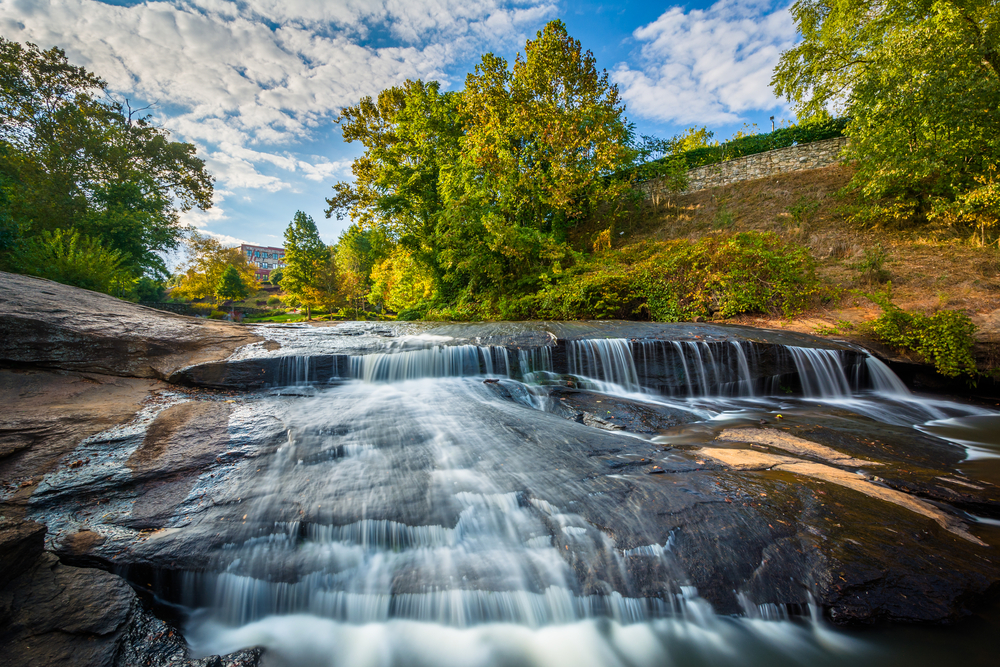 A low rise waterfall over wide granite rocks with bright green trees on the surrounding banks on a sunny day; Falls Park on the Reedy is on the top things to do in Greenville SC.