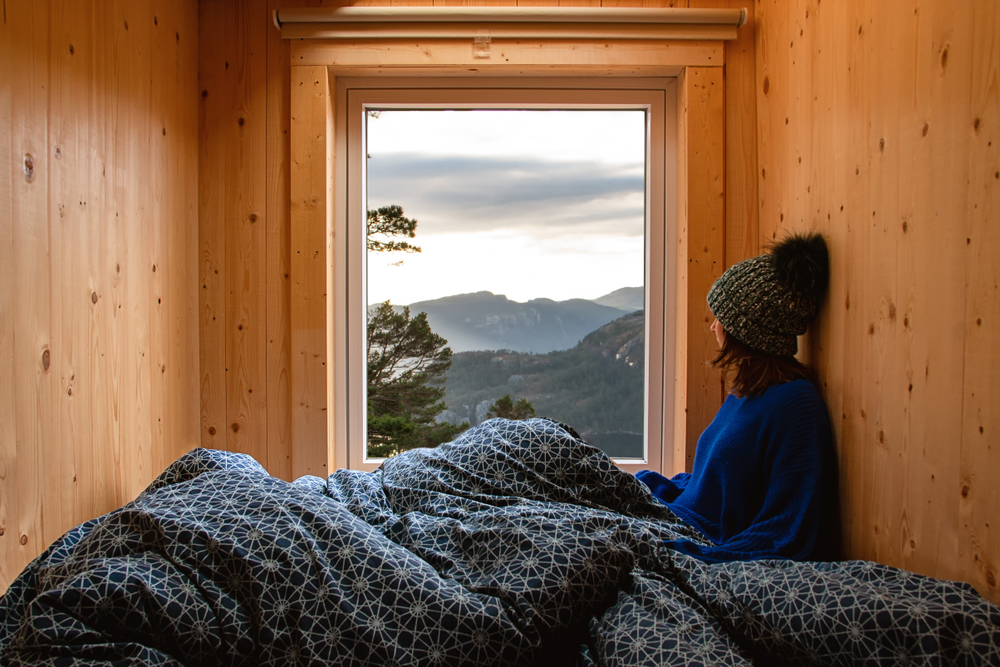 A woman sitting in a rustic cabin wrapped in a blanket looking out the window at rolling mountains. 