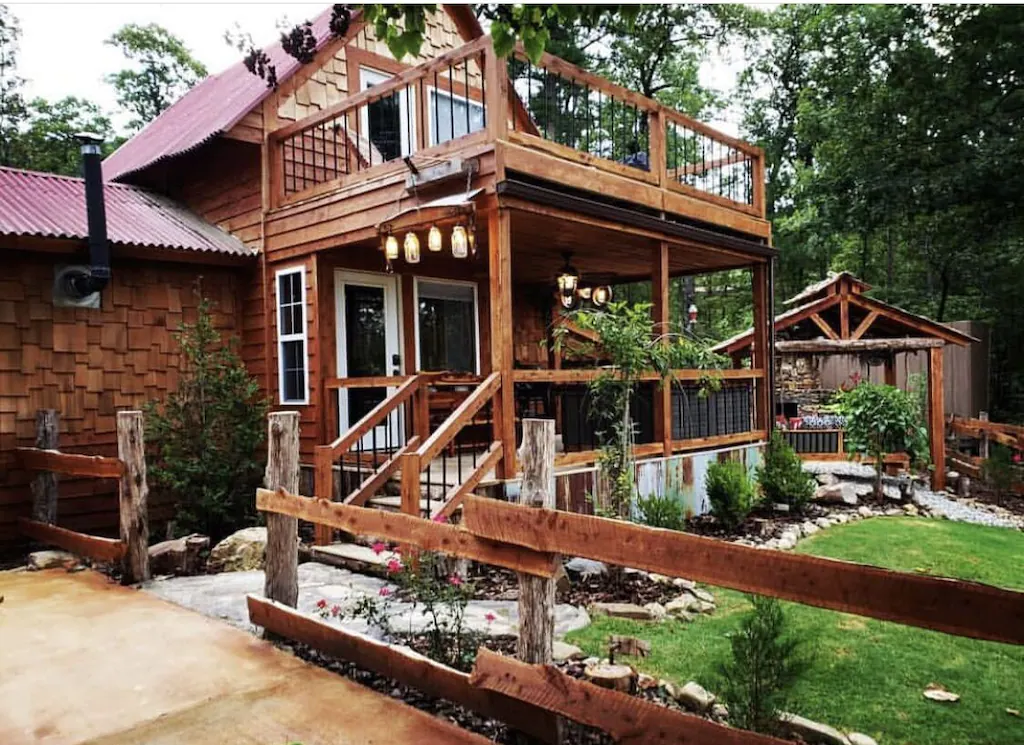 A two storey wooden cabin with wood trim and a wood fence. A patch of grass to the right and tall green trees surrounding. 