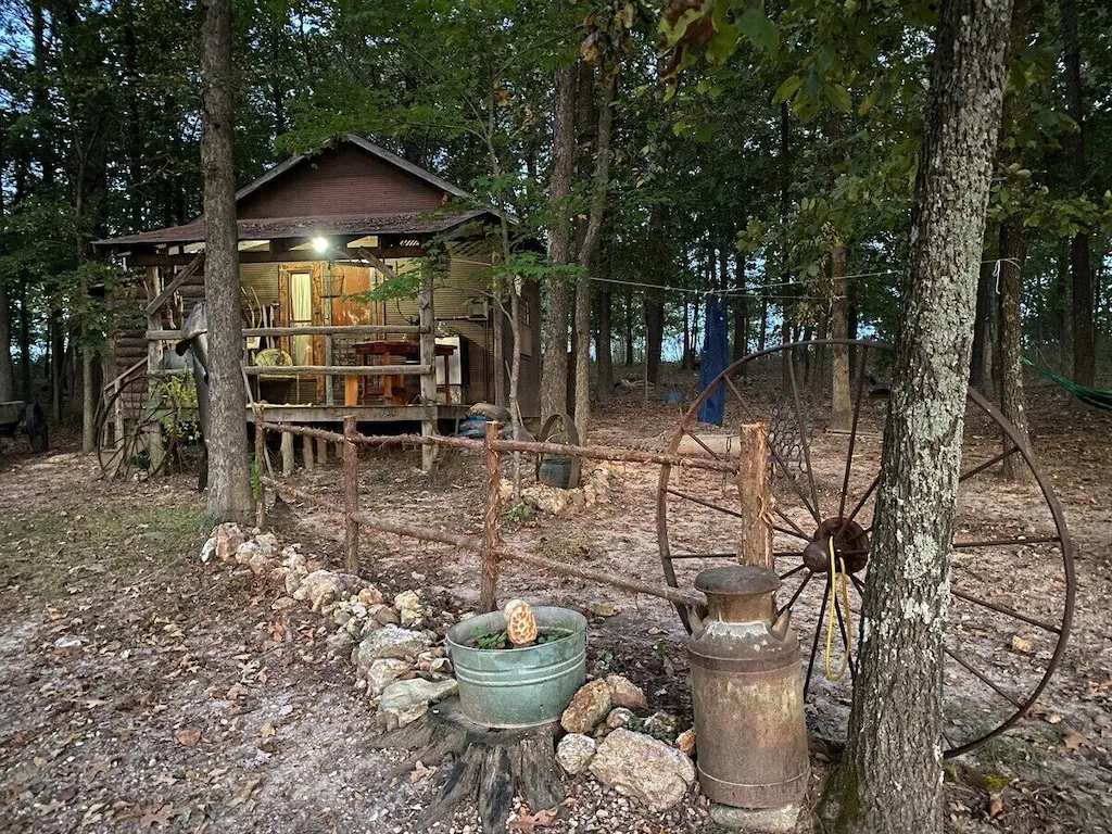 A large rusted old wagon wheel in the foreground, behind it a cabin set amongst a forest. 