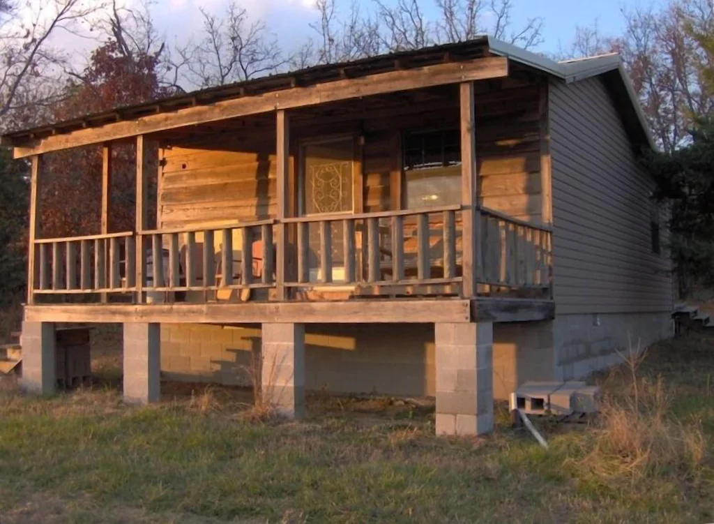 Long shadows across a close up of a grey wooden cabin on raised concrete. There's a porch with a log balustrade.