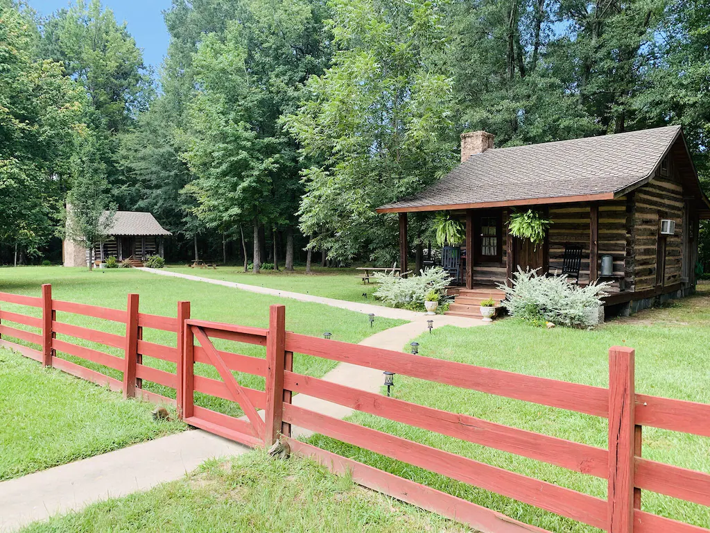 Two wooden cabins sit behind a red wooden fence among grass in front of a row of green trees for glamping in Arkansas