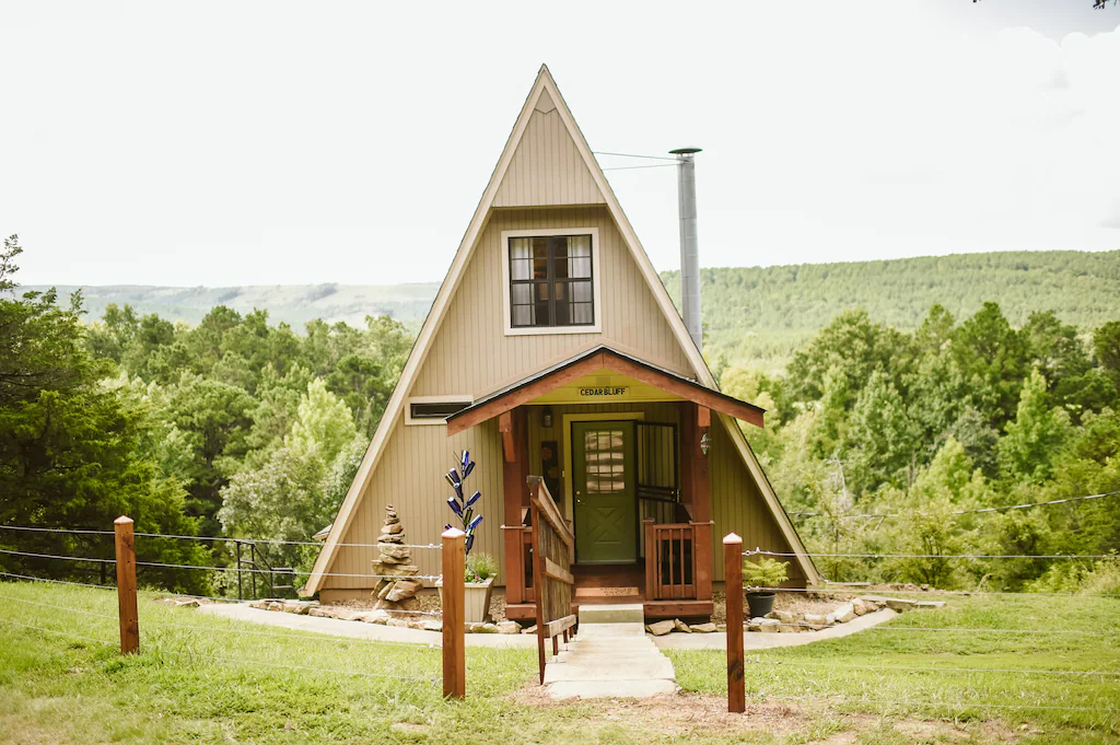 An A-Frame cabin sits on green grass in front of a low lying green forest glamping in Arkansas