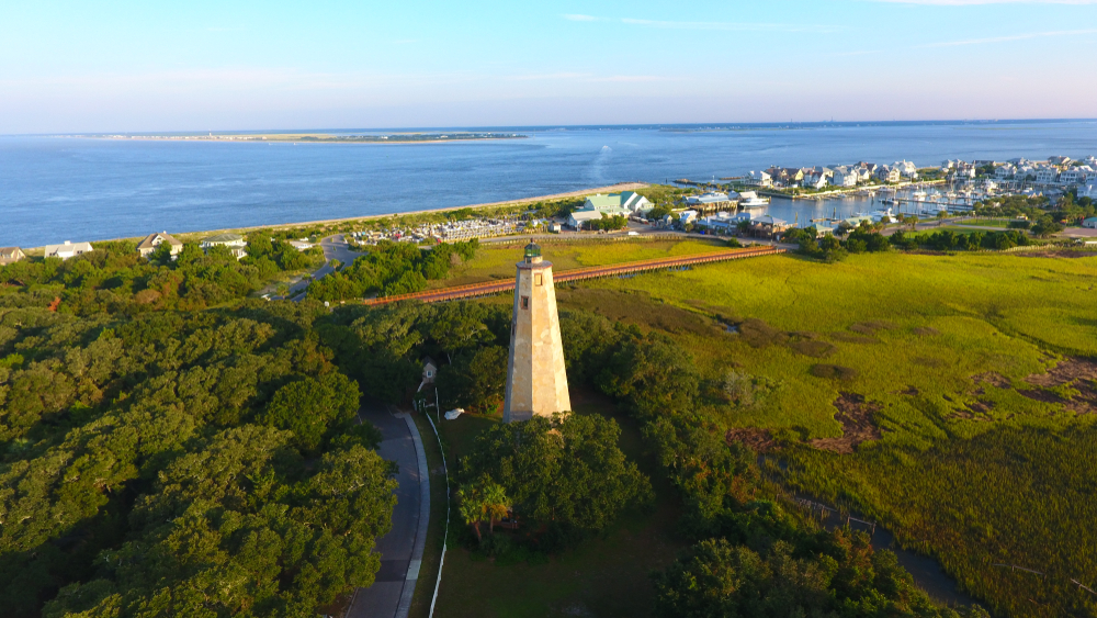 Photo taken from the air showing a forest and paddock, a marina in the distance and the ocean on the horizon. There is a lighthouse in the foreground. 