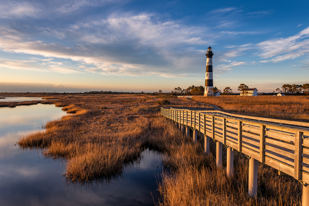A black and white lighthouse in the middle distance, a wooden boardwalk leads up to it. Surrounding it is marshland and blue sky. 