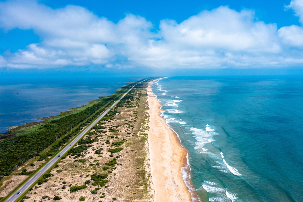 An aerial photos of an island strip with ocean on both sides and a road going through the center. The island goes to the horizon and the sky is blue with puffy white clouds.