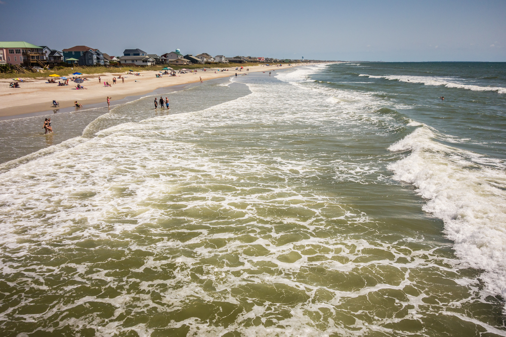 Waves and sea foam crash on the shore of the beach, the photo shows the beach all the way into the distance. There are people on the shore and houses to the left. 