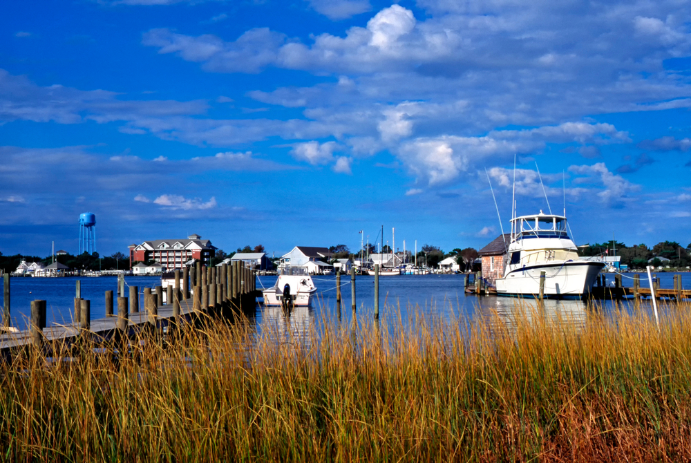 Tall grass in the foreground of a harbour with boats tethered to two piers. Buildings in the distance and a blue sky on the islands in north carolina