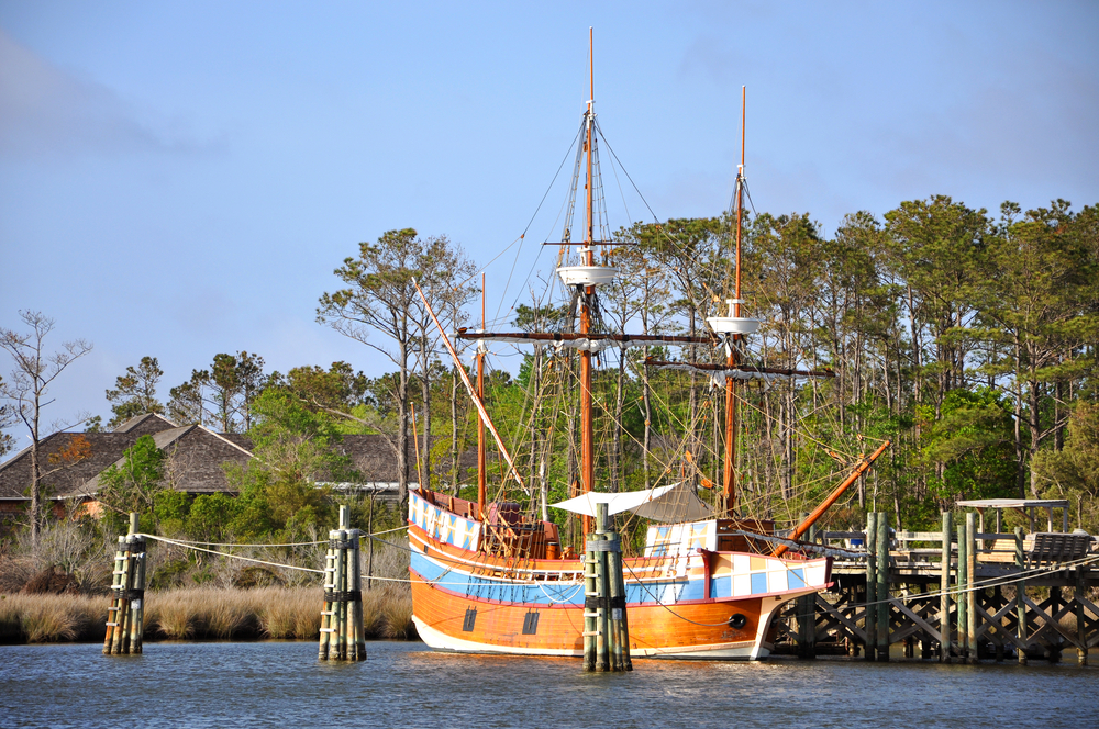 A wooden sailing ship docked at a small pier with trees in the distance. 