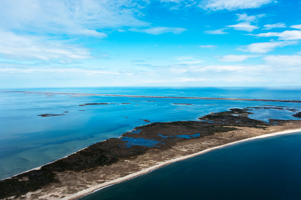 An aerial photo of a narrow barrier island surrounded by blue sky and sea. 