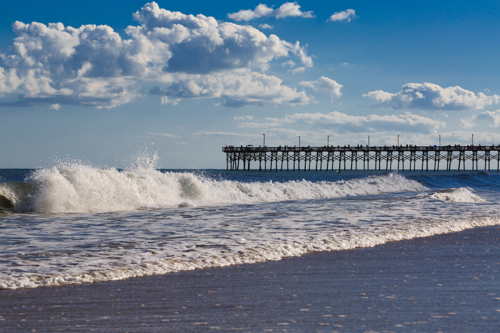 Strong waves crash on the shore of a beach A tall wooden pier is in the middle distance. Blue skies and puffy white clouds on a north carolina island