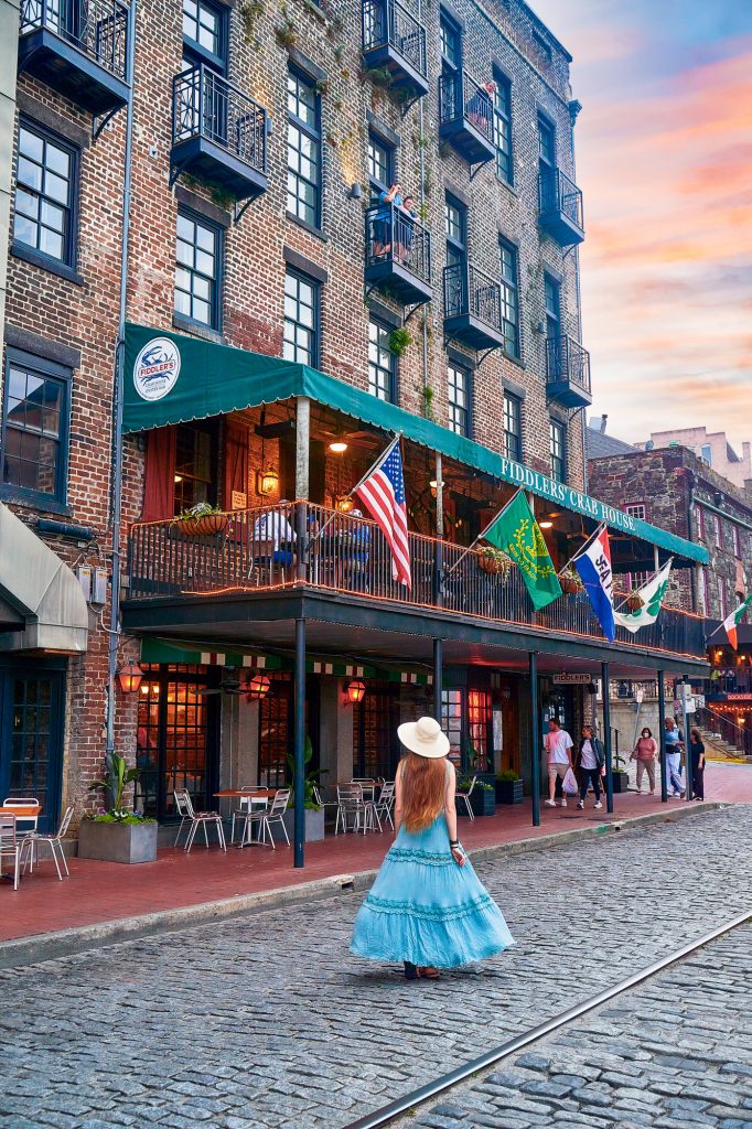 A woman in a long blue dress walks by the awning-covered balcony of the brick-walled Fiddlers' Crab House, where people can stop on a food tour as one of the best things to do in Savannah.