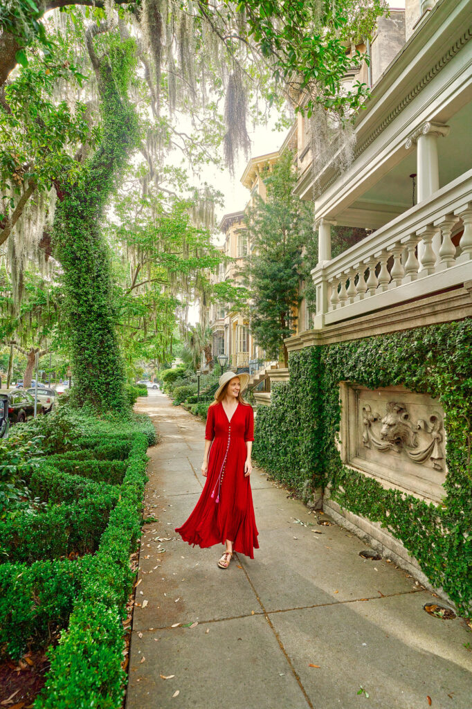 A woman in a long red dress and sunhat walks down a sidewalk in Savannah, GA flanked by staircases and moss-covered trees. Going on a walking tour is one of the best things to do in Savannah.
