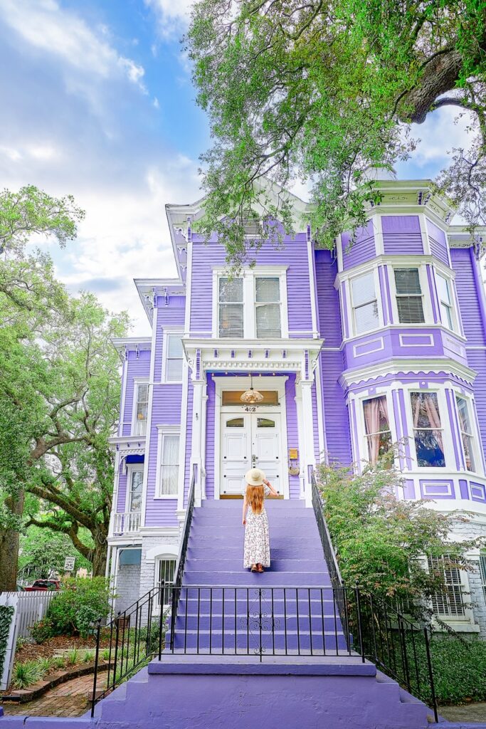 A woman with long hair in a floral dress stands on the steps of a light purple Victorian house on Gaston Street, where admiring the old homes is one of the best things to do in Savannah.