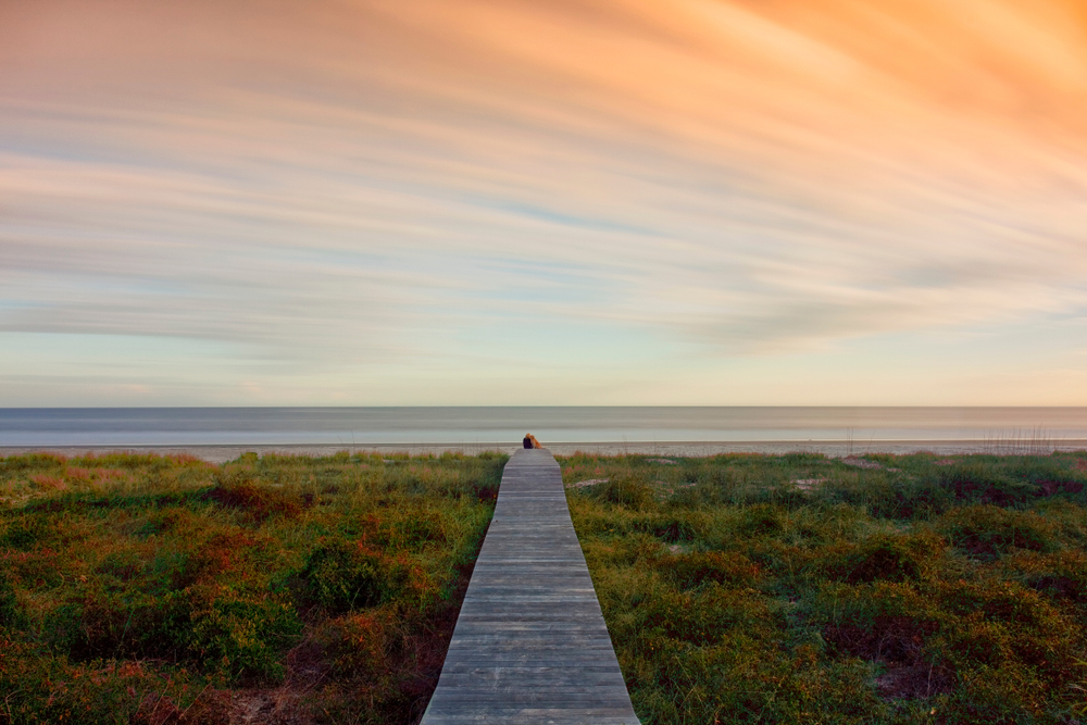 A long wooden boardwalk that goes over grassy dunes towards a calm and pristine beach at sunset. You can just barely see two people sitting on the very end of the boardwalk.  