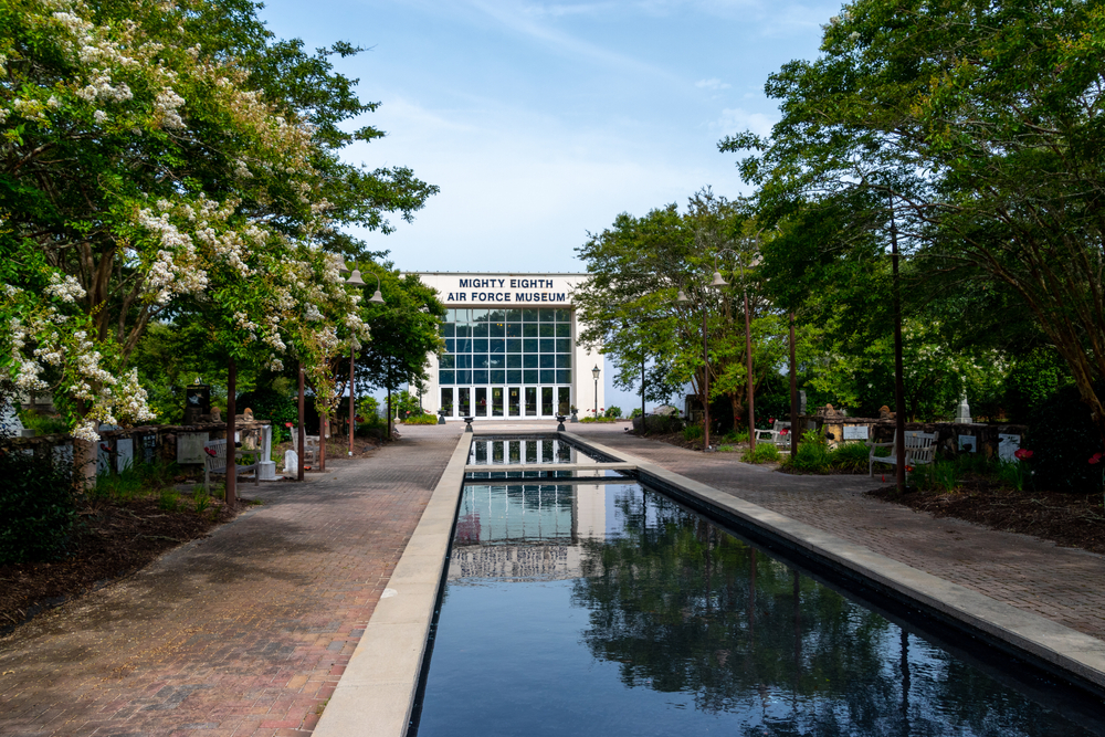 The front of the Mighty Eighth Air Force Museum in Savannah GA. It is a large white building with windows all along the front. In front of the building is a large water pool. 