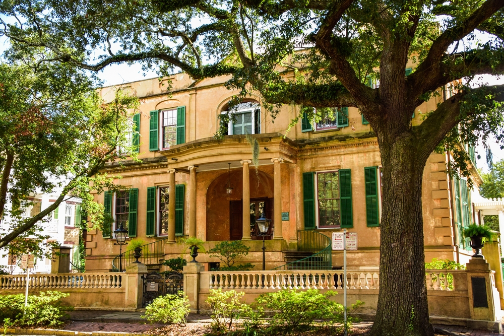 The front exterior of a historic mansion house museum in Savannah. It is yellow with green shutters and is surrounded by trees and shrubs. 