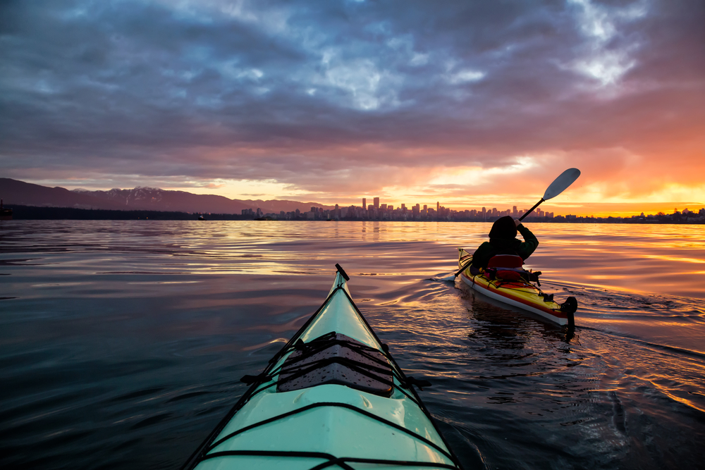 The view of the front of a person's kayak, and another person ahead of them kayaking, in the ocean with a city and trees in the distance while the sun is setting. 