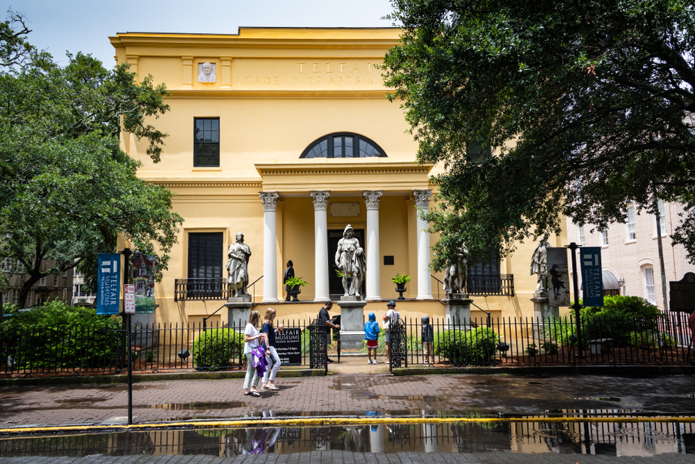 The front of a Greek revival mansion that is painted buttercream yellow. It has sculptures and trees in front of it. 