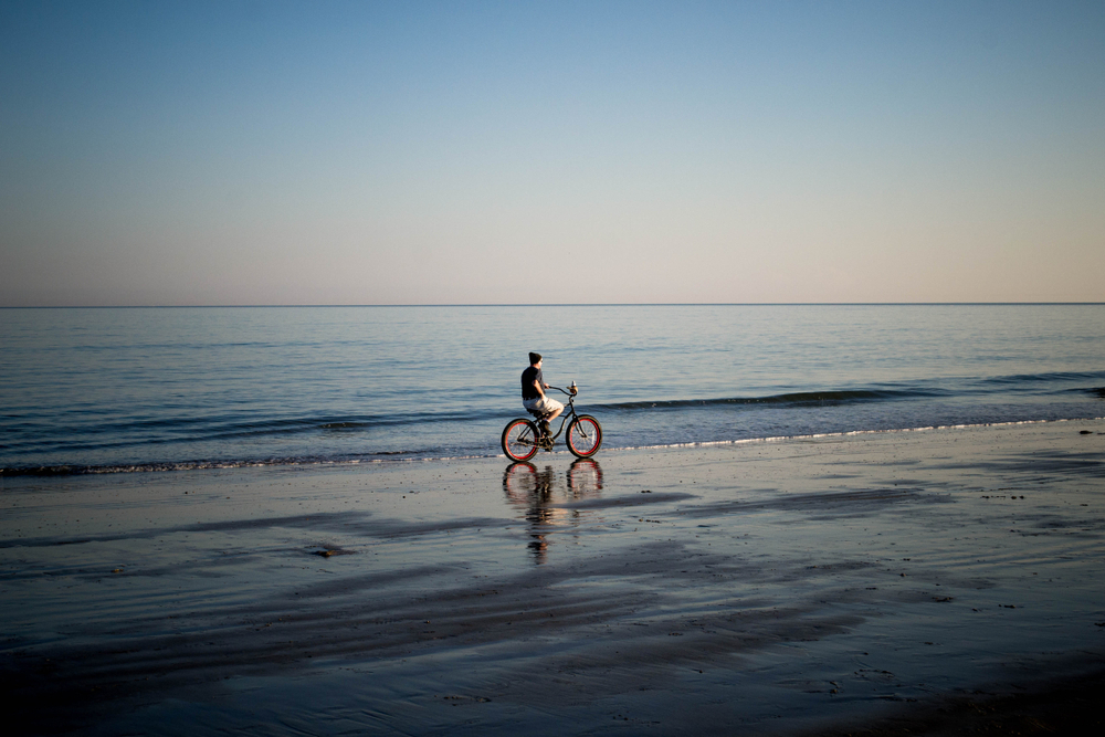 A male riding a bike on the beach. The sea is calm and the beach is deserted. 