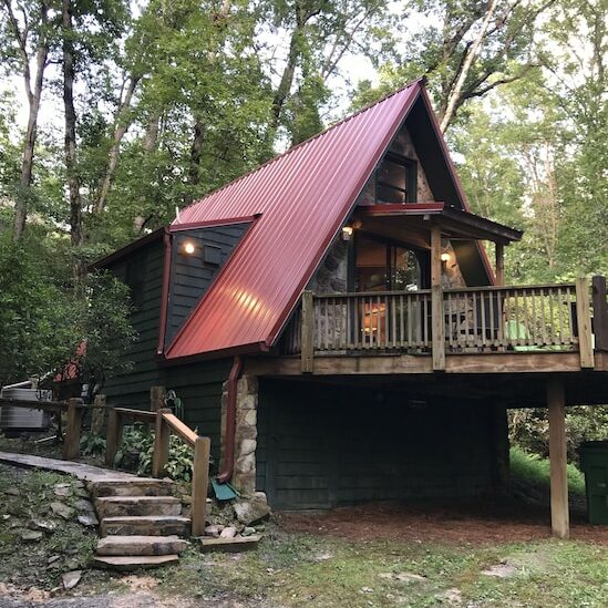 A cabin in the woods with a peaked red roof, deck and wooden walk way with lots of green trees. 