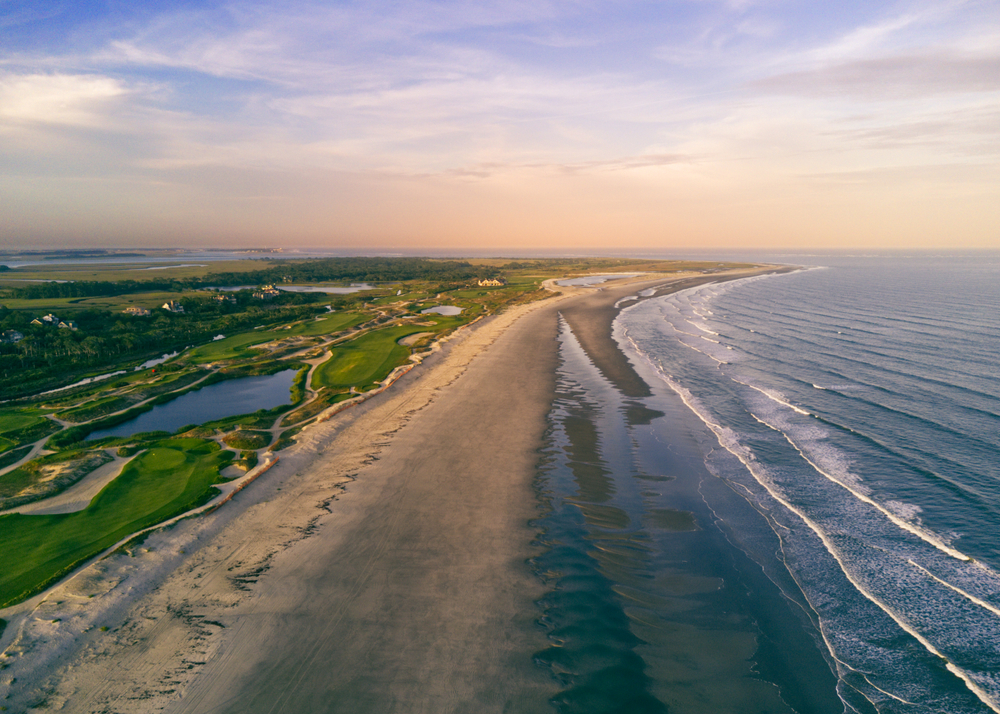 A overhead view of Kiawah Island, which is one of the beach towns in South Carolina, shows the coast in the soft evening, where waves crash against sand, and well-manicured golf courses sit to the left.