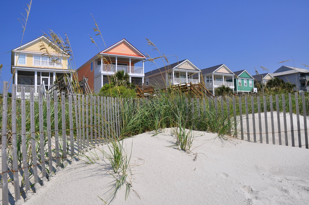 At one of the beach towns in South Carolina, two story homes sit in pastel colors, overlooking the sand dunes and fences on the shore.