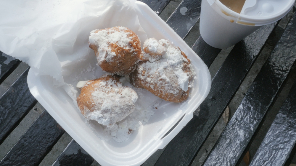 Three beignets sit in a to go container, fried and topped with powdered sugar. These are great snacks but also perfect for breakfast in New Orleans! 