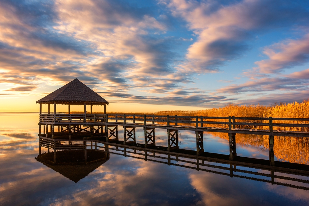 Calm water, sunset reflections, and gazebo over the water with grass in the background. 