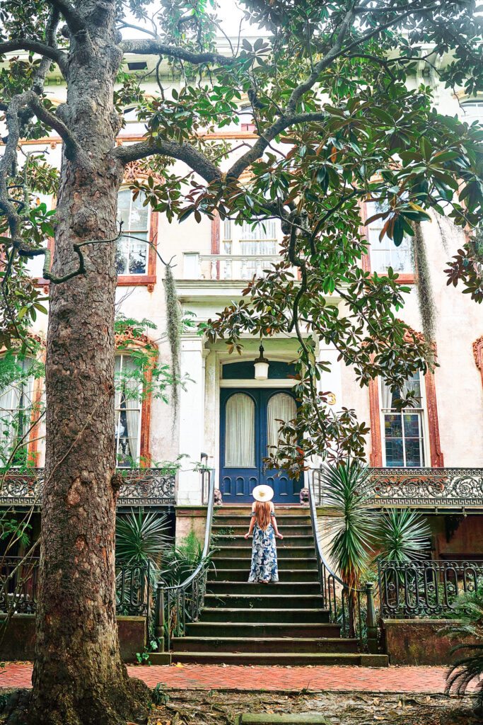 A woman with long hair, wearing a sundress and straw hat, stands on the steps of a mansion in the historic district, where you can find many boutique hotels in Savannah.