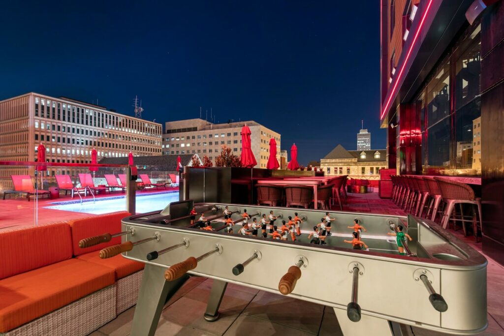 A rooftop terrace bar with chairs, a bar a swimming pool and a foortball game in the foreground. 