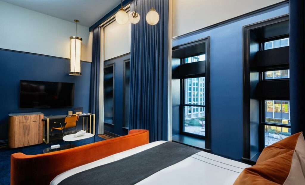 A hotel room decorate in dark blue, rust orange, and white. It has a bed, tv, art deco lights, and large windows. 
