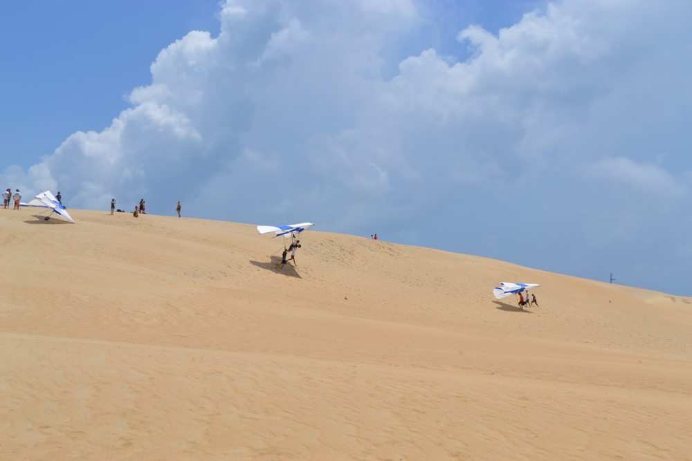 sand dunes with people hang gliding off of them in the jockeys ridge state park, one of the best things to do in kitty hawk 