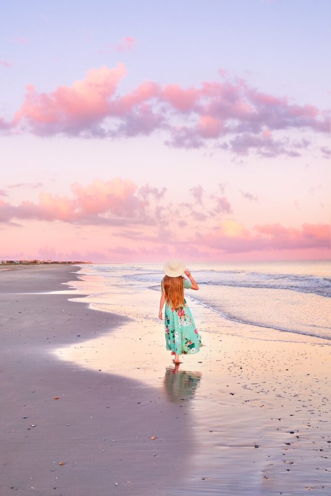 Woman in a floral dress and sun hat walks along a beach during a pastel sunset.