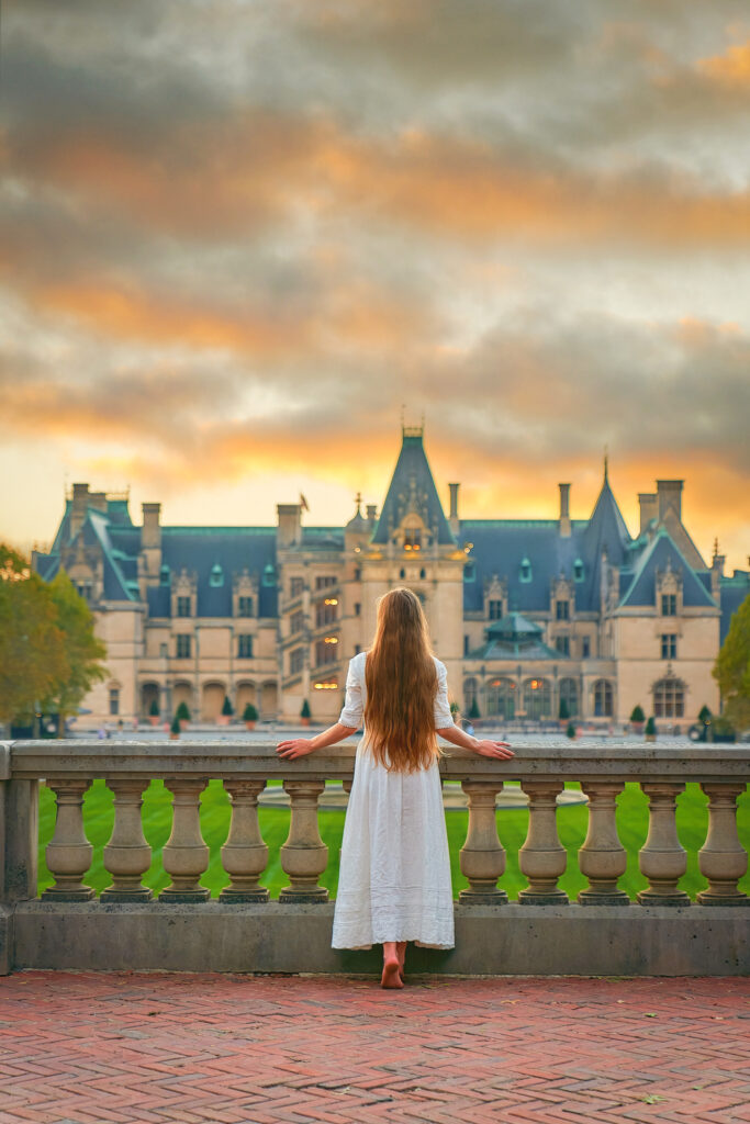 Woman in a white dress stand on a terrace overlooking the Biltmore Estate at sunset in North Carolina.
