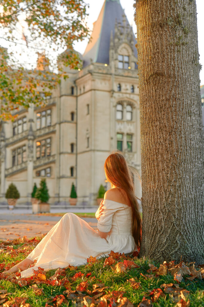 Woman in a white dress sits at the base of a tree in fall leaves looking at the Biltmore Estate.