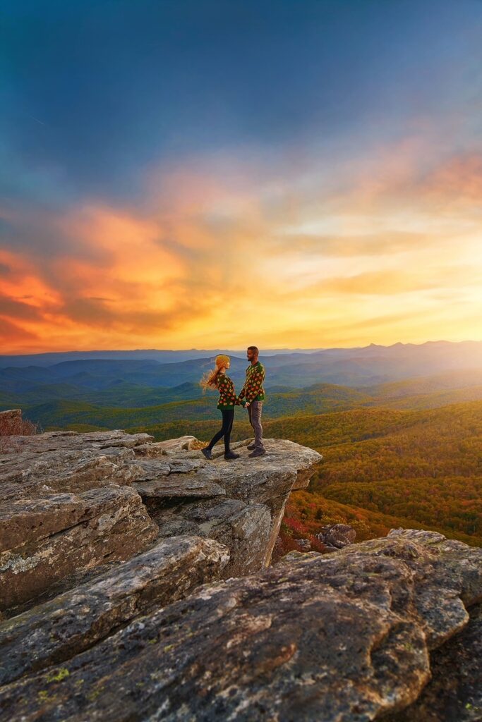 A couple in matching fall sweaters  stands on a rocky ledge overlooking fall mountains in North Carolina.