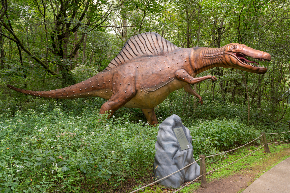 A dinosaur statue stands next to a path in a forest.